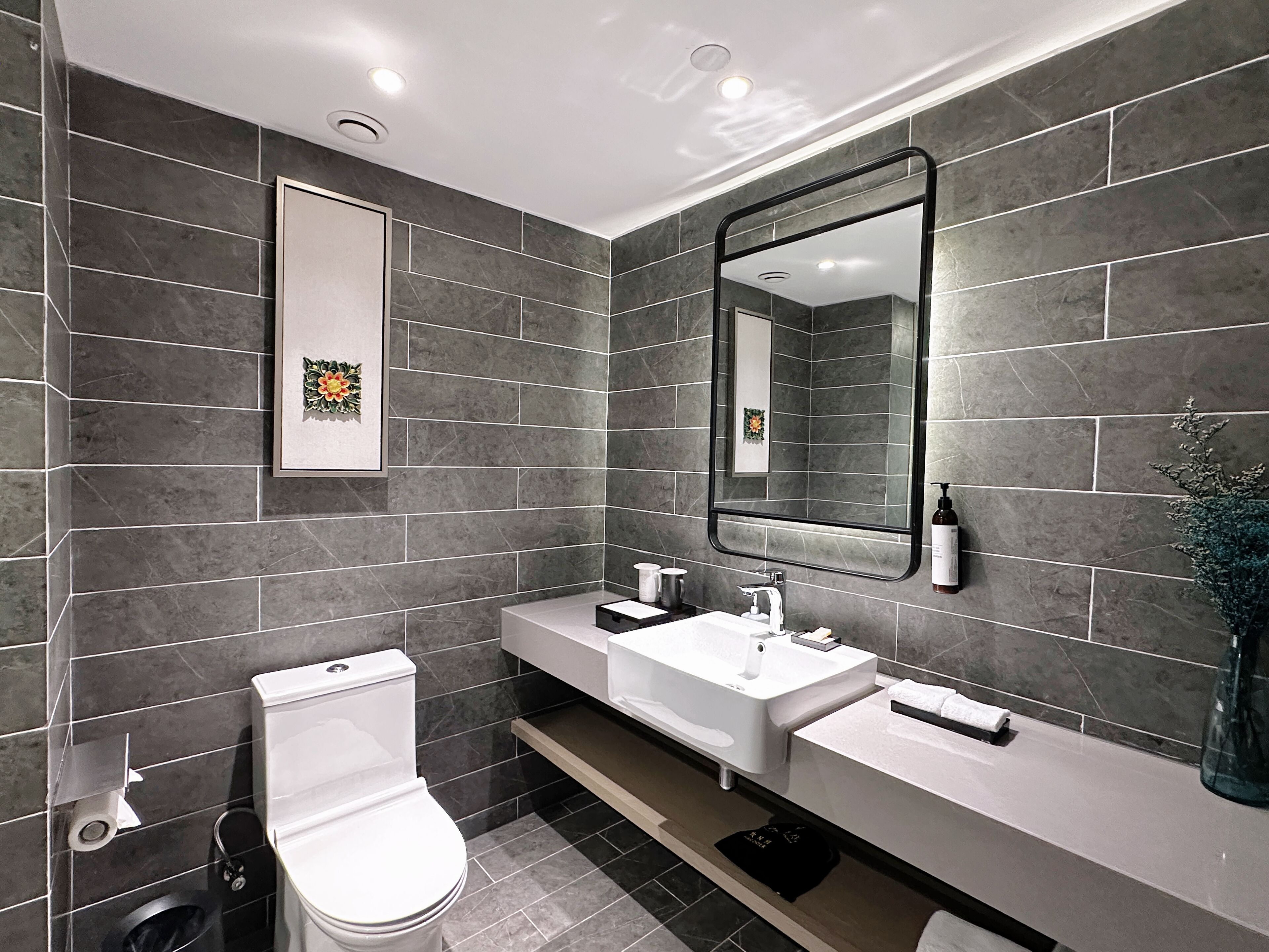 Un baño moderno con azulejos grises oscuros, un inodoro blanco, un lavabo blanco y un espejo grande.