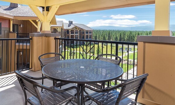 Un balcon avec une table ronde et des chaises, donnant sur une zone boisée et un bâtiment à la façade jaune.