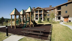 A playground with wooden play structures and a picnic table in a landscaped area with a building in the background. - Luxurious 1 bedroom condo at the entrance to Yellowstone NP. (West Yellowstone)
