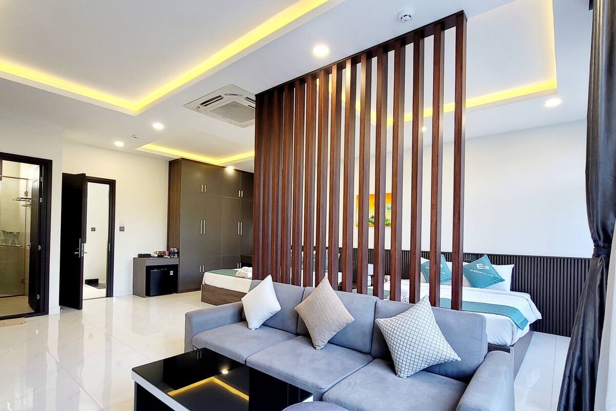 Une chambre d’hôtel moderne avec un canapé gris, une table basse noire, une cloison en bois et un lit avec des oreillers.