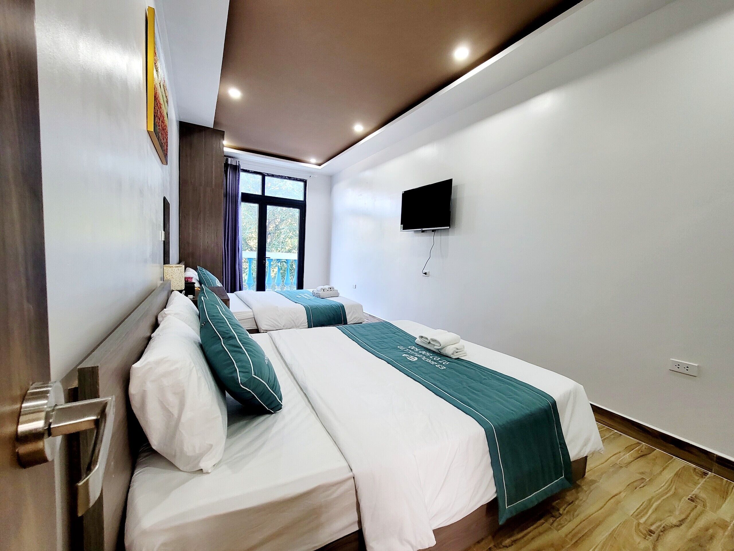 A hotel room with two beds, a TV mounted on the wall, and a window with a view of greenery.