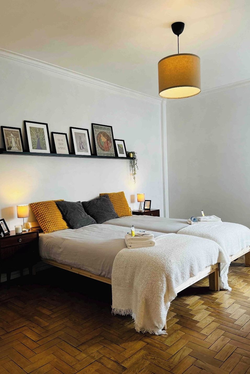 Ein Hotelzimmer mit zwei Betten, einem Holzboden im Fischgrätmuster, einer hängenden Leuchte und einem an der Wand befestigten Regal mit gerahmten Bildern.