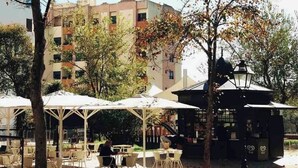 An outdoor seating area with tables and umbrellas, surrounded by trees and a building in the background. - Lisbon - Low Cost - Airport - Subway (Lisboa)