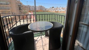 A balcony with a round table and two wicker chairs, overlooking a sports field and residential buildings.