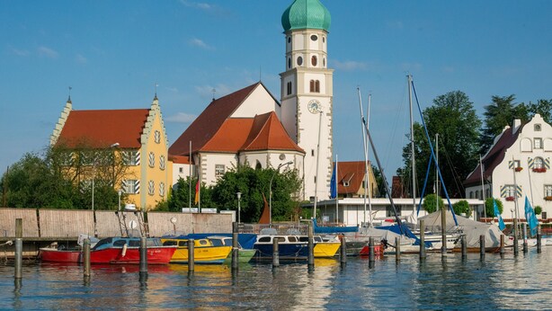 A waterfront with boats docked, a church with a green dome, and traditional buildings. - Apartment "Haus Kaulitzki" with Lake View, Private Terrace & Wi-Fi (Wasserburg (Bodensee))
