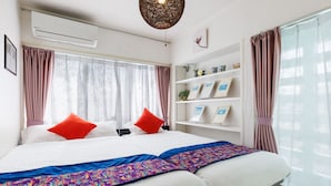 Una habitación de hotel con una cama grande, dos almohadas, una colcha colorida, una estantería con fotografías enmarcadas y una ventana con cortinas.