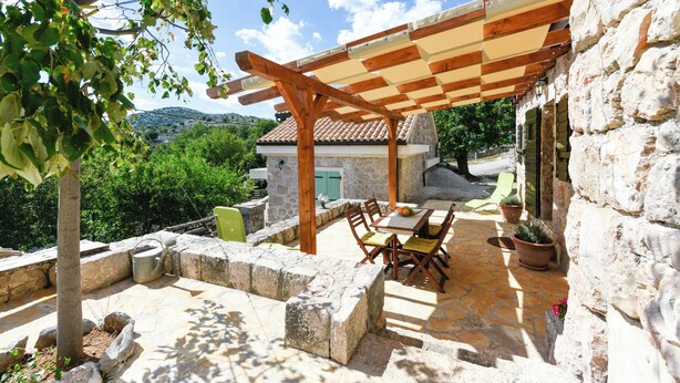 Een terras buiten met stenen vloer, houten meubilair en een pergola.