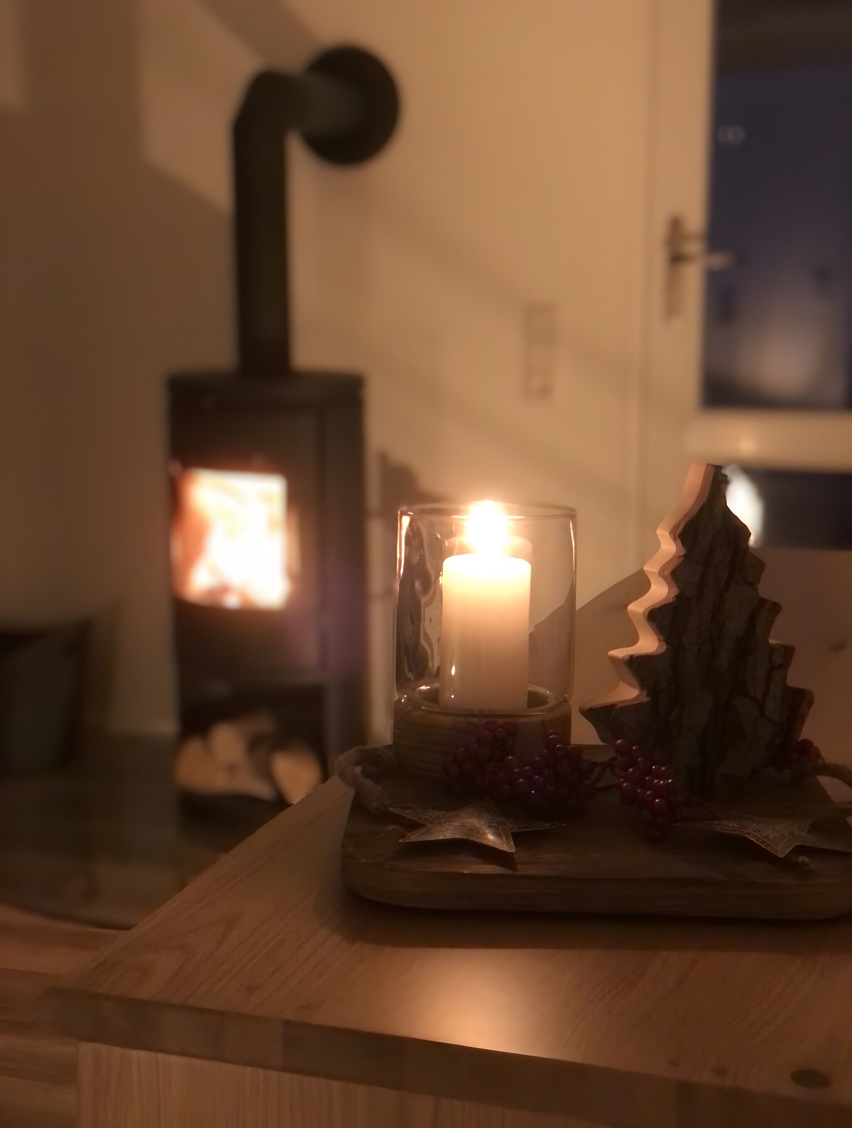 A candle in a glass holder on a wooden table with a rustic decoration, a fireplace in the background, and a window.