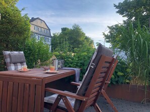 An outdoor patio with a wooden table, two chairs, a cushion, a cup, a bowl, and a kettle.