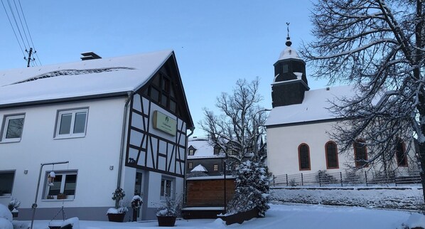 A traditional half-timbered building with a sign, a church with a steeple, and snow-covered surroundings.