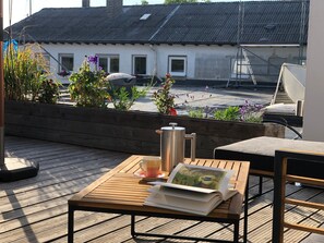 Eine Holzterrasse mit Tisch, Stuhl, Buch, Kaffeekanne und Fenster mit Blick auf ein Gebäude mit einem Metall-Dach.
