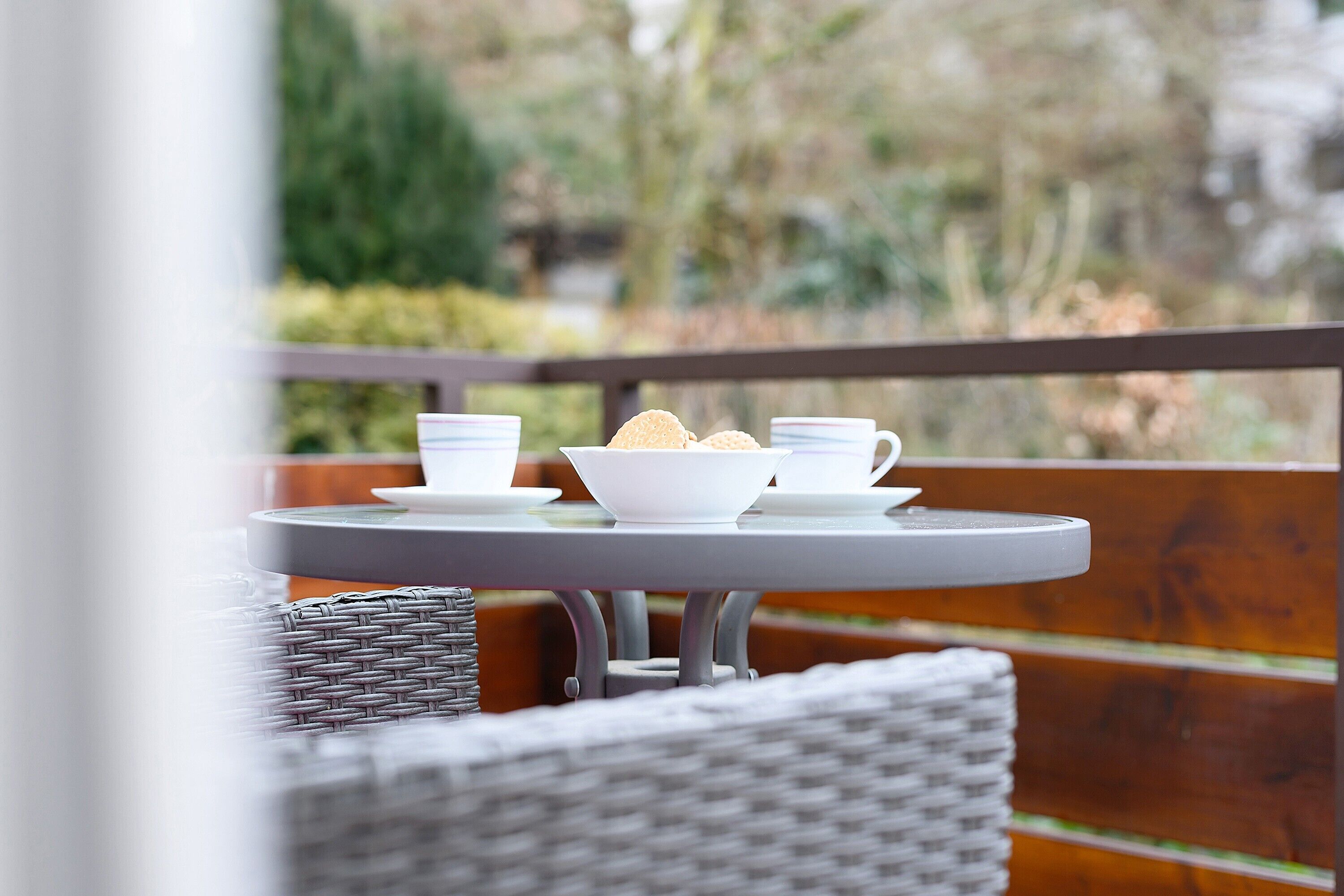 Un balcon avec une table dressée pour deux personnes, deux tasses blanches, un bol blanc et une pile d’assiettes blanches.