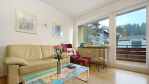 Un salon avec un canapé beige, une table basse au motif coloré, un fauteuil rose et une vue sur un balcon doté d’une balustrade.