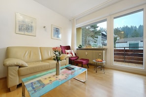 A living room with a beige sofa, a coffee table with a colorful design, a pink chair, and a view of a balcony with a railing.