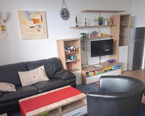 En stue med en sort lædersofa, en rød skammel, et træbord, en sort lænestol, et fjernsyn, en reol og et køleskab.