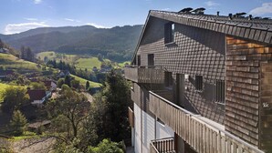 Un immeuble à plusieurs étages, doté d’un toit en pente, de balcons en bois et offrant une vue sur une vallée parsemée de maisons et de montagnes.