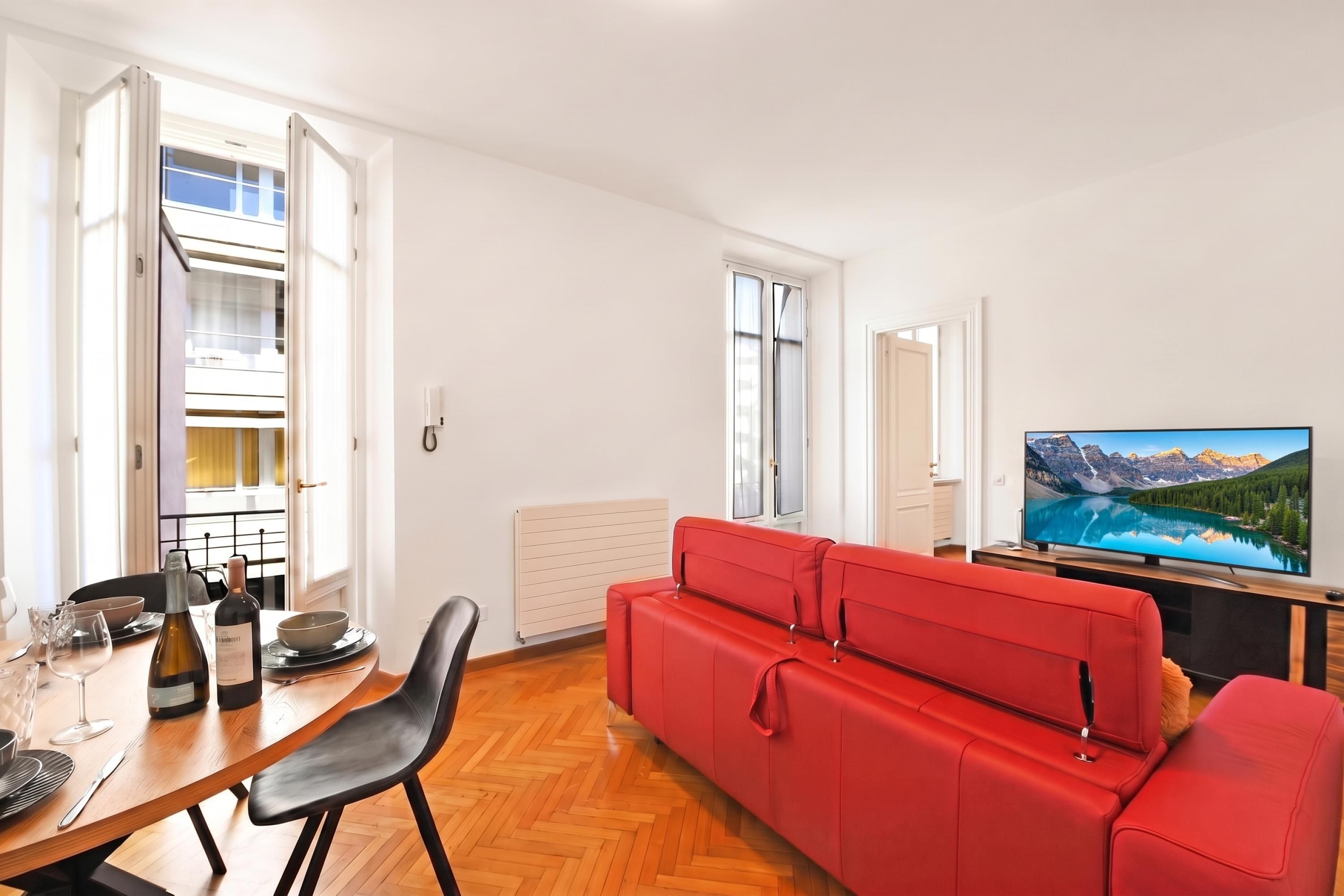 Uma sala de estar moderna com um sofá vermelho, uma mesa de jantar com cadeiras, uma televisão e vista para montanhas pela janela.