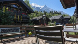 Una terraza de madera con un banco y una mesa, vistas a la montaña y un edificio con un letrero.