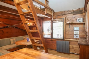 Interno di una baita in legno con letto a soppalco, una scala in legno, un divano, un tavolo da pranzo e una finestra con persiane.