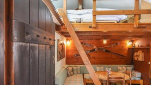 Interior acogedor de una cabaña con una cama en el altillo, una zona de comedor con una mesa y sillas, y un rincón de descanso con cojines.
