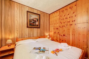 A wooden-paneled room with a bed, a nightstand with a lamp, a framed picture, and a wooden wardrobe.