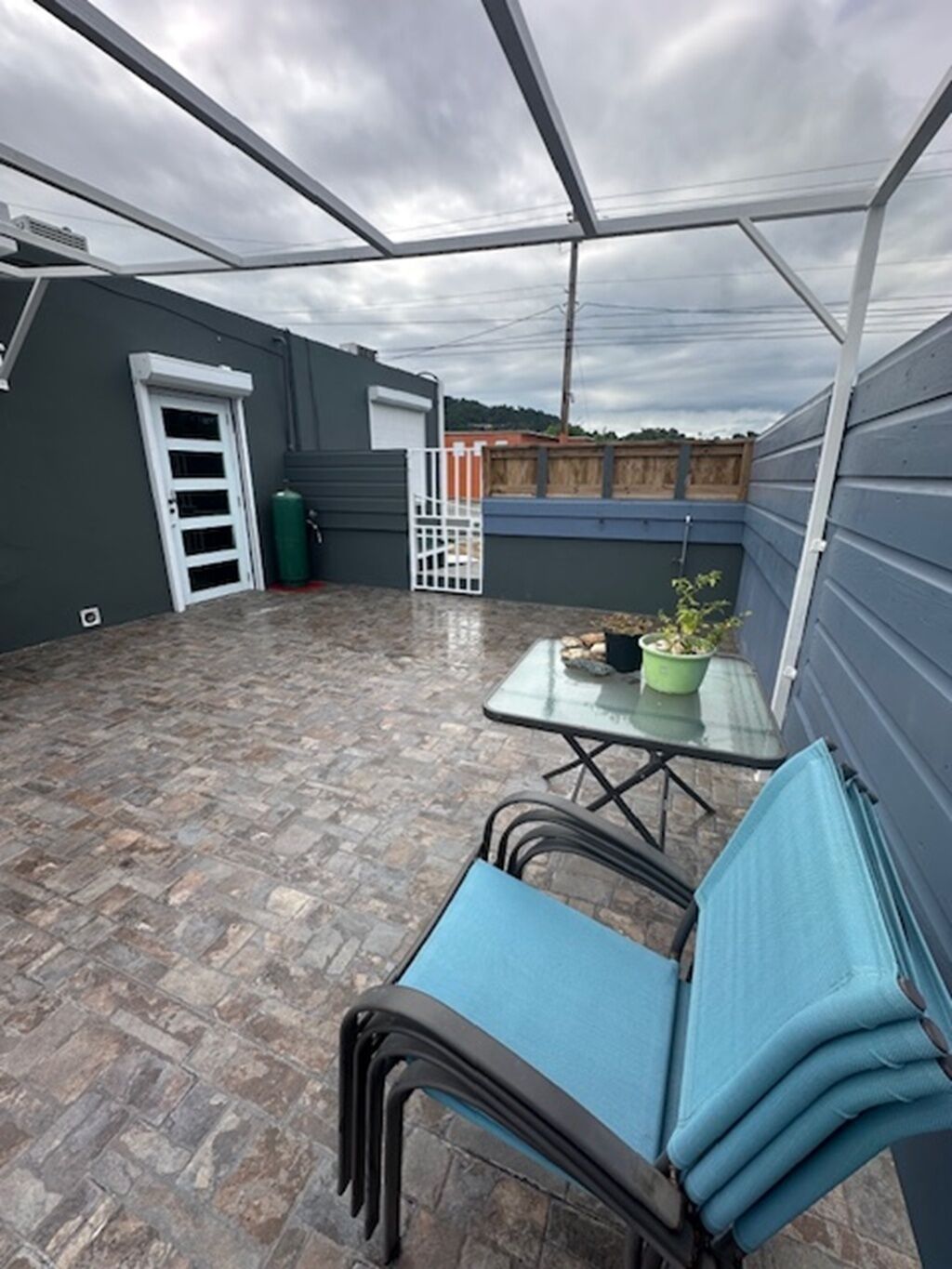 Een overdekt terras met een tafel, stoelen, een deur en een groene afvalbak.