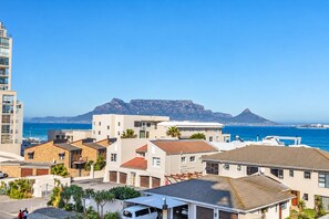 A coastal residential area with modern buildings, a clear blue sky, and a prominent mountain in the background.