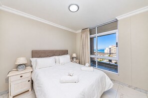 A hotel room with a large bed, a nightstand, a lamp, a window with a view of buildings and the sea.