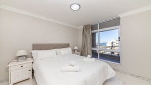 A hotel room with a large bed, a nightstand with a lamp, a window with a view of buildings, and a ceiling light. - Modern Blouberg Beach Pad | views pool parking (Bloubergrant)