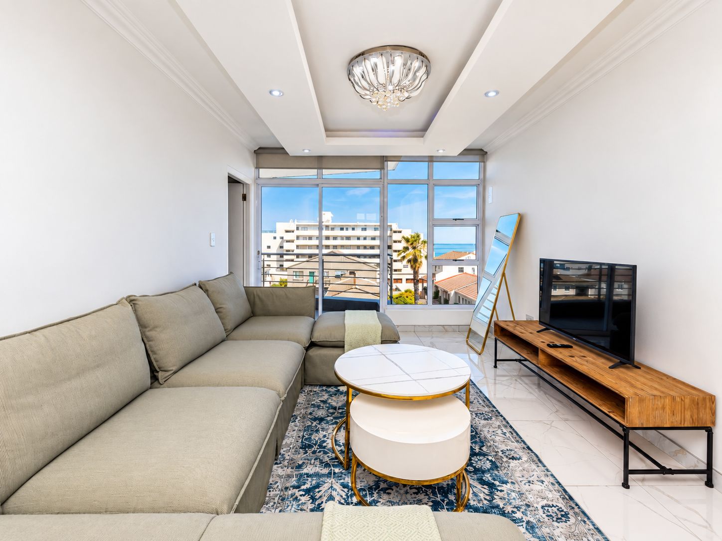 A modern living room with a sectional sofa, a round coffee table, a TV stand, and a large window offering a view of buildings and palm trees.