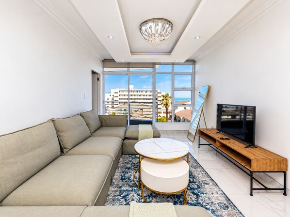A modern living room with a sectional sofa, a round coffee table, a TV stand, and a large window offering a view of buildings and palm trees.
