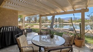 A covered patio with a round table and chairs, a grill, and a view of a lake and desert landscape. - Peaceful Waterfront Getaway (pet-friendly) (Palm Desert)