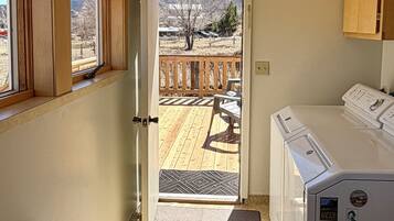 A room with a wooden bench, a washing machine, a door leading to a balcony with a view of a mountainous landscape.