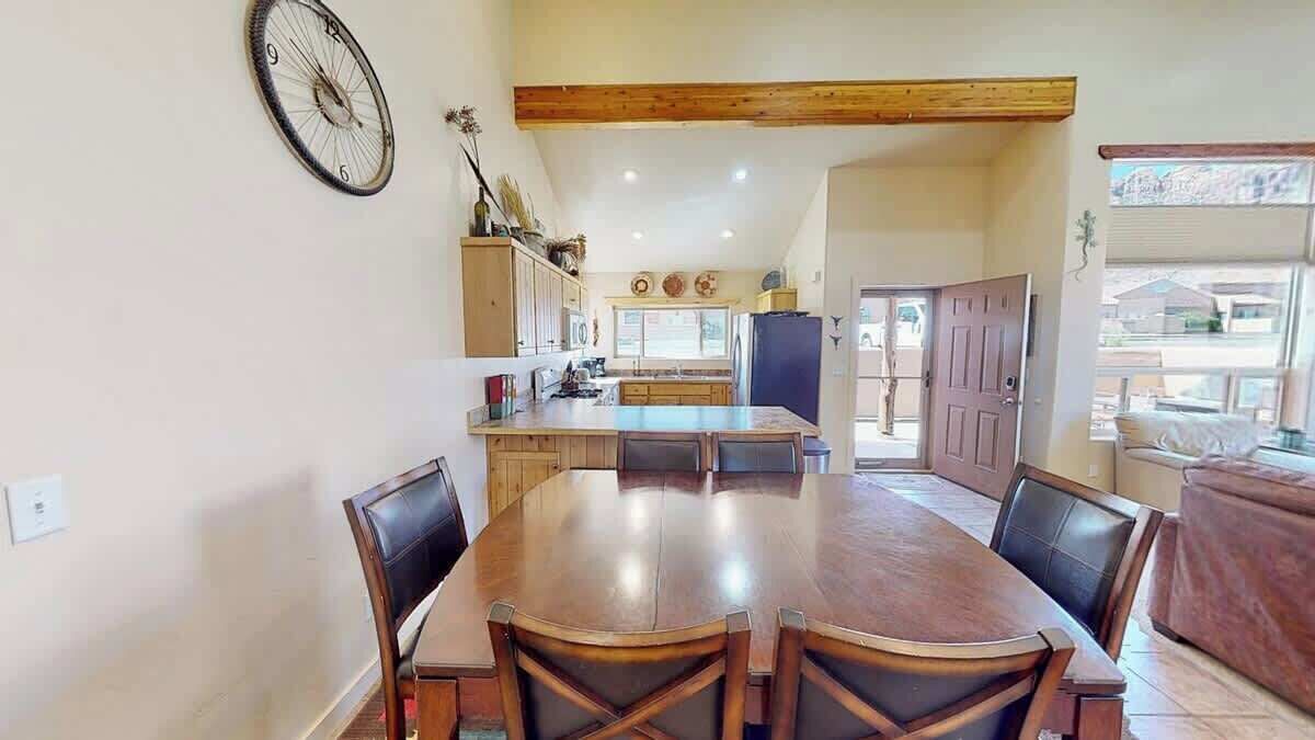 A dining area with a large wooden table and chairs, a kitchen in the background, and a door leading outside.