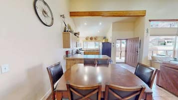 A dining area with a large wooden table and chairs, a kitchen in the background, and a door leading outside.