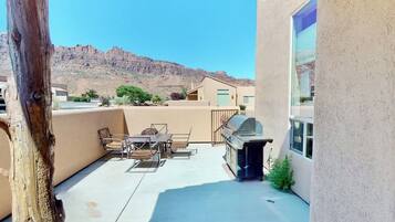 An outdoor patio with a barbecue grill, a table with chairs, and a view of a mountain range.