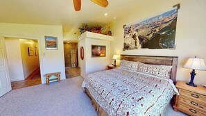 Um quarto com uma cama grande, uma mesa de cabeceira, um banco, um ventilador de teto e um quadro na parede.