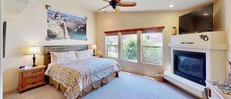 A bedroom with a bed, bedside tables, a ceiling fan, a fireplace, a TV, and a window with a view.