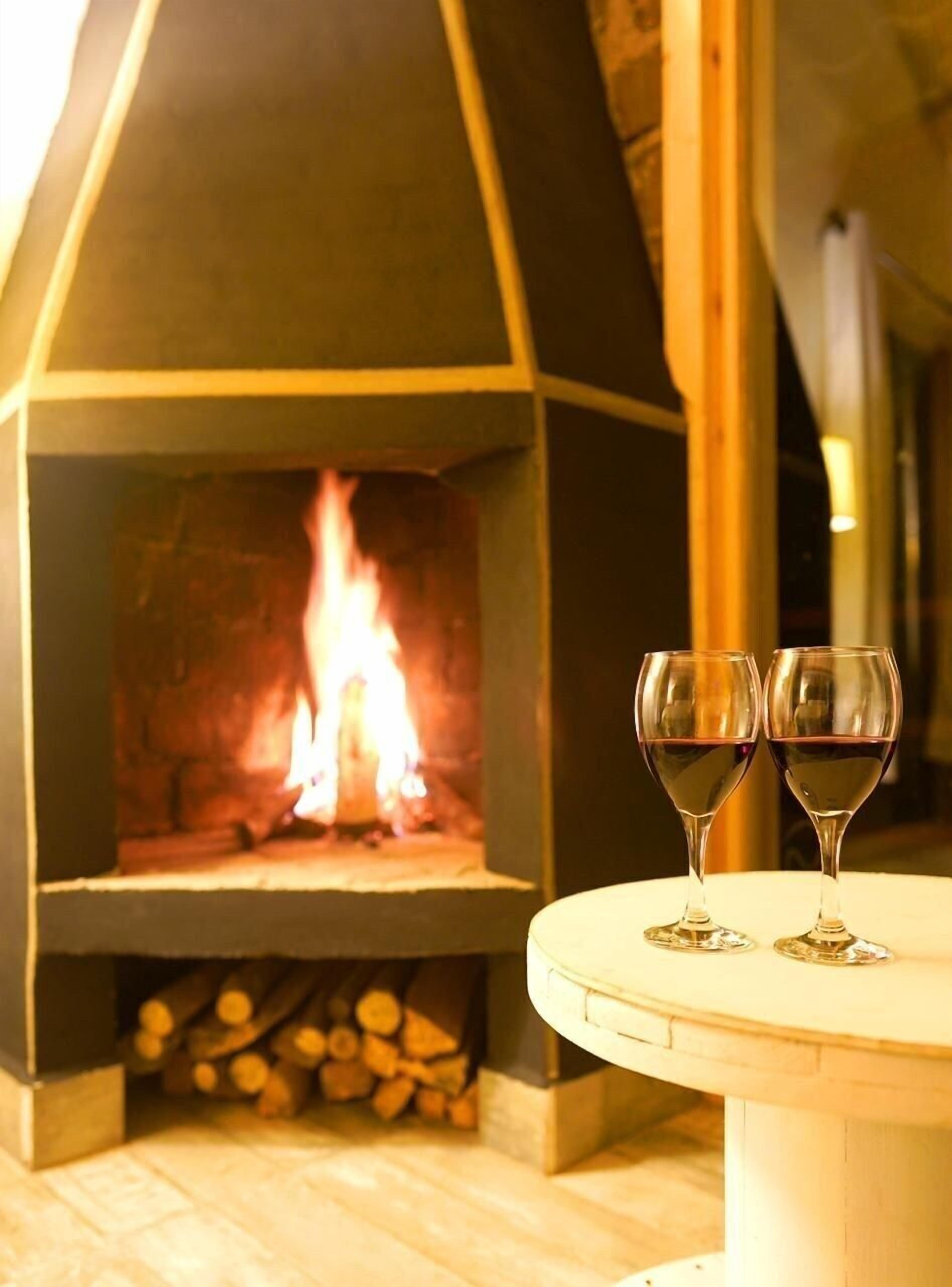 A fireplace with a visible fire, two wine glasses on a table, and a stack of firewood in the background.