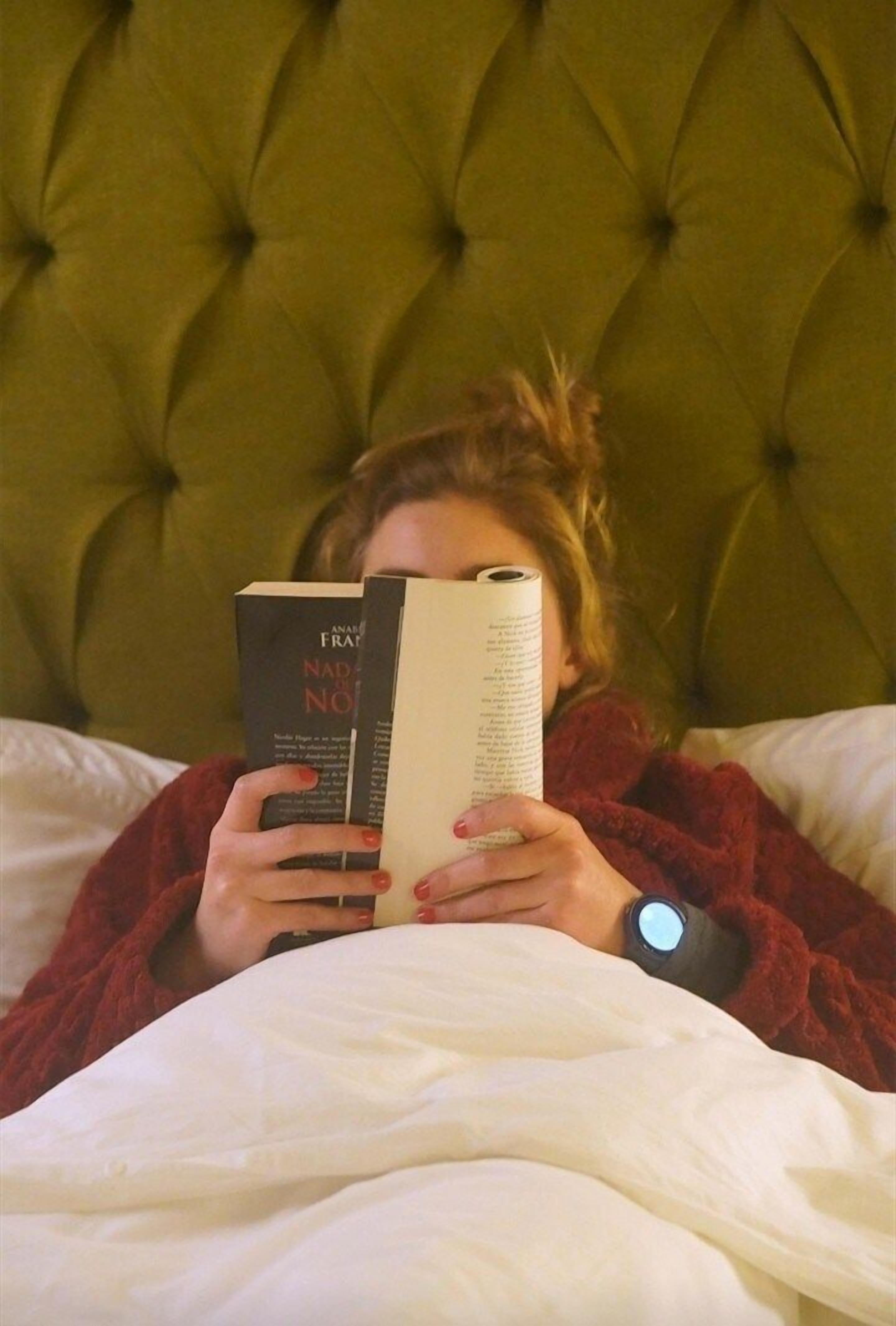 Una persona recostada en una cama con un cabecero tapizado en verde, leyendo un libro.