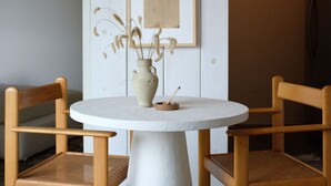 Ein runder Tisch mit weißem Fuß, begleitet von zwei hölzernen Stühlen mit Flechtbespannung. Auf dem Tisch steht eine Vase mit getrockneten Pflanzen.