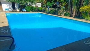 Piscina com superfície de água azul cristalina, rodeada por um deck de concreto e algumas cadeiras de descanso.
