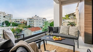 Un balcon avec un canapé, une table sur laquelle est disposée une assiette de fruits, et une vue sur le paysage urbain.
