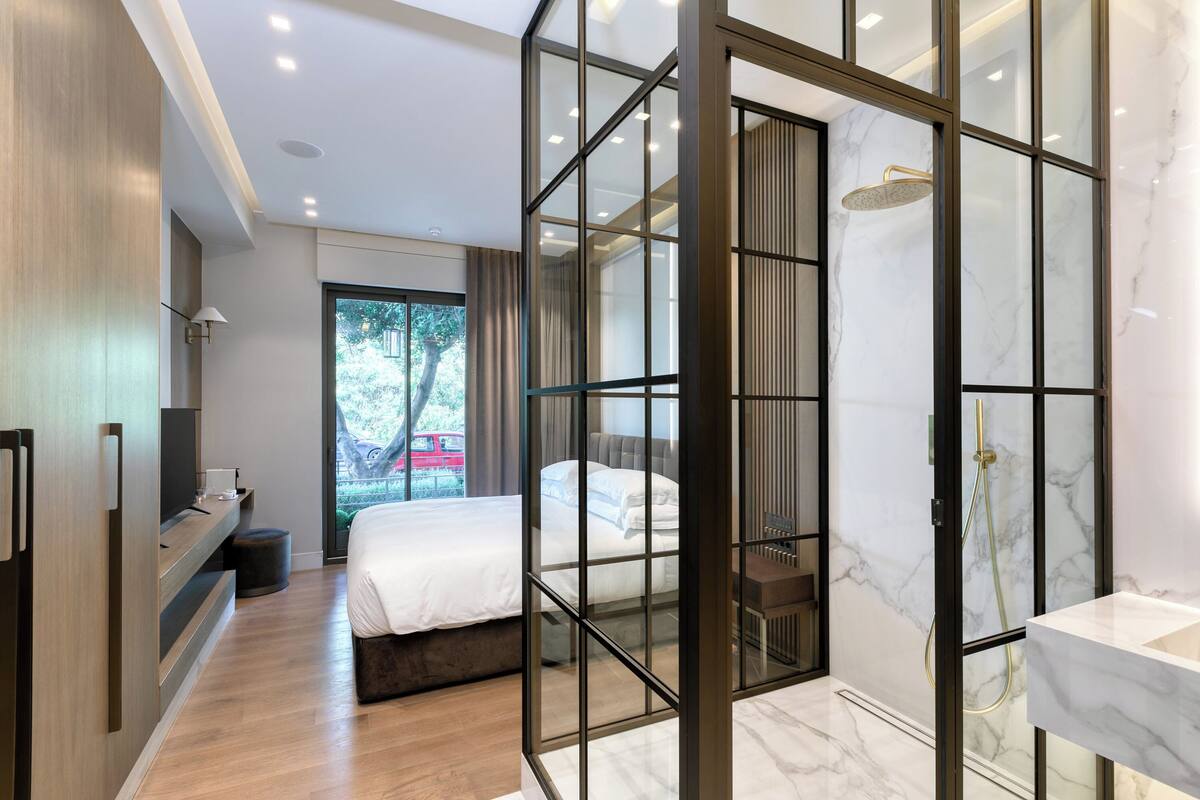 A modern hotel room with a large bed, a walk-in closet, a bathroom with a glass shower enclosure, and a view of the outdoors.