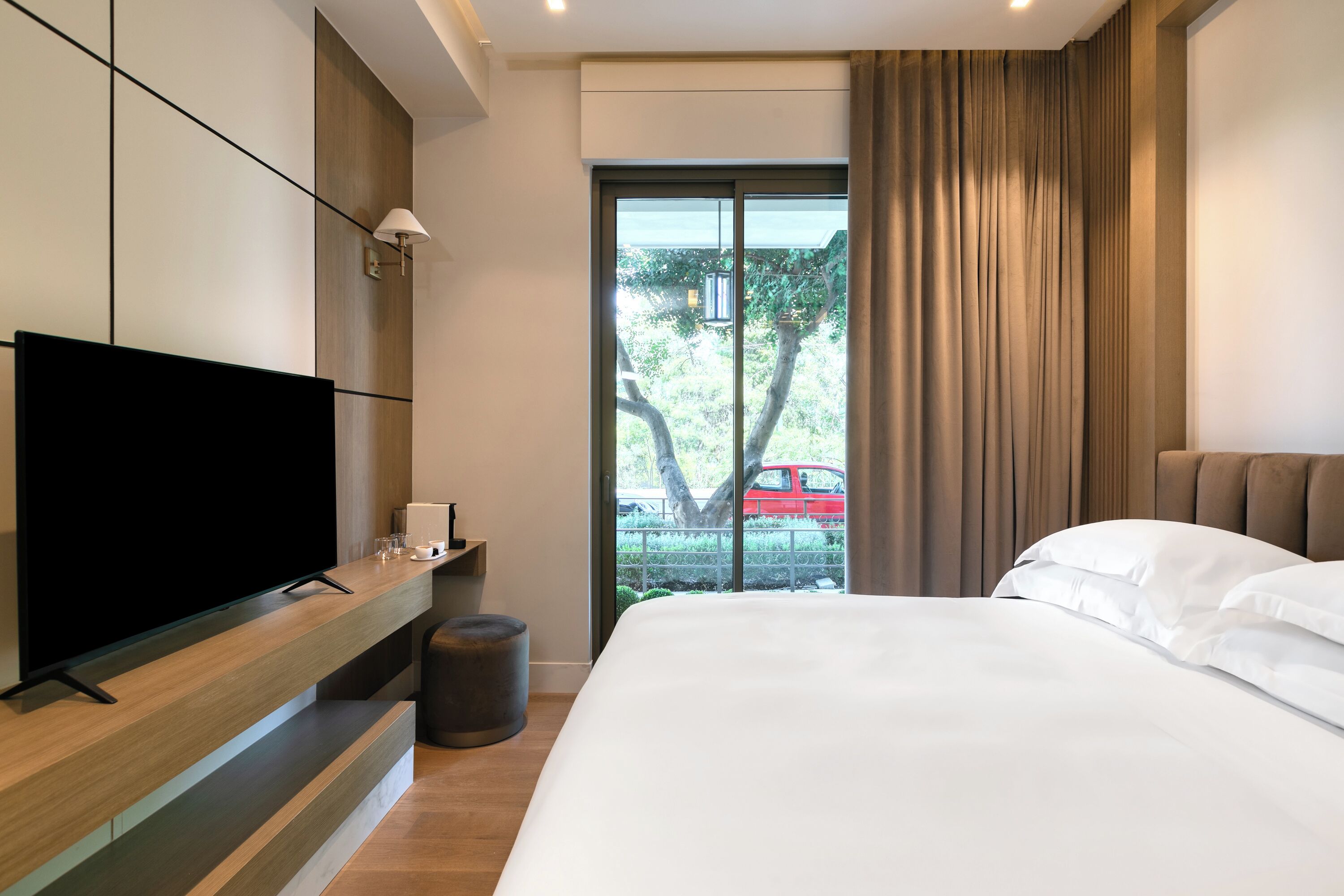 Ein modernes Hotelzimmer mit einem großen Bett, einem Flachbildfernseher auf einer Holzkonsole, einem kleinen Tisch mit Lampe und Blick auf ein rotes Auto durch ein großes Fenster.