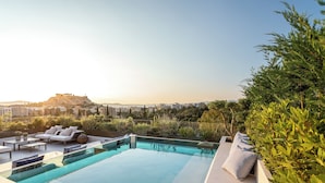Una piscina infinita en la azotea con asientos exteriores, rodeada de vegetación y vistas a un paisaje urbano.