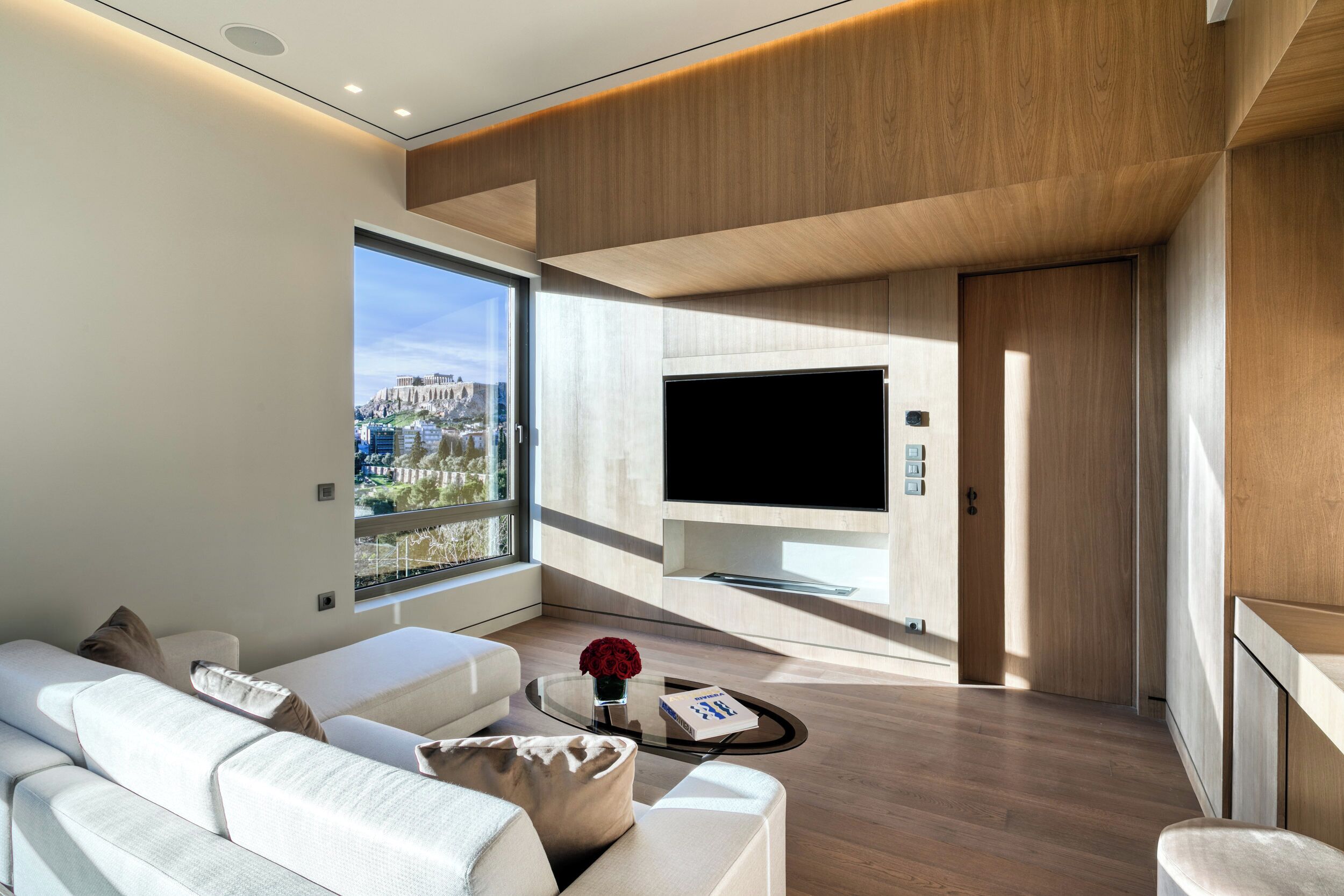 A modern living room with a large TV, comfortable seating, a coffee table with a vase, and a view of the city.