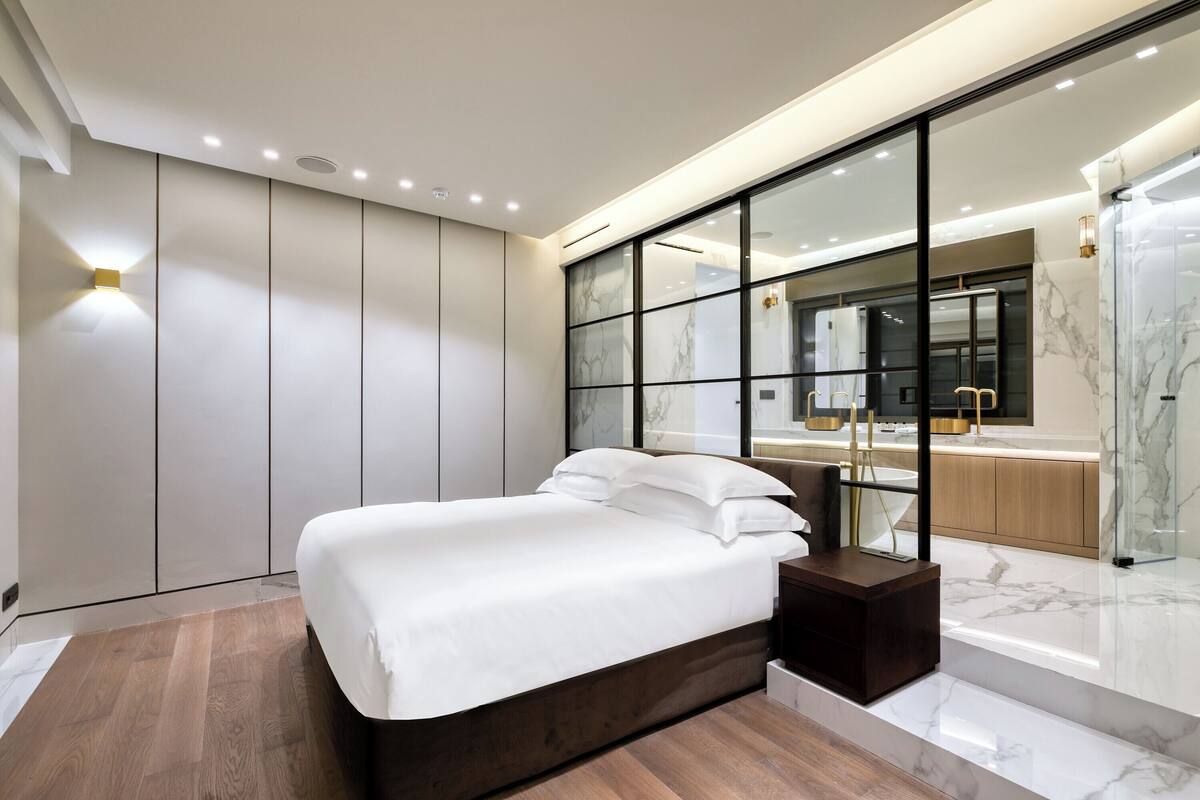 A modern bedroom with a large bed, a wooden nightstand, a glass partition, and a bathroom with a marble countertop.