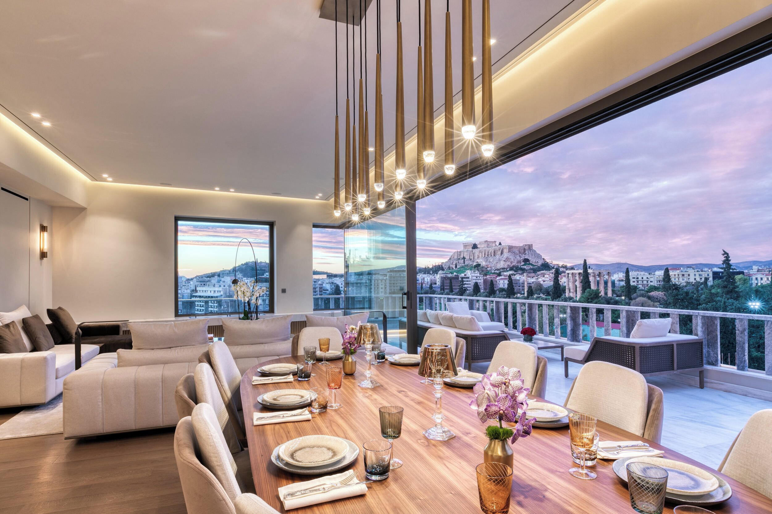 A modern dining area with a wooden table set for six, large windows offering a city view, and a cozy seating area with sofas.