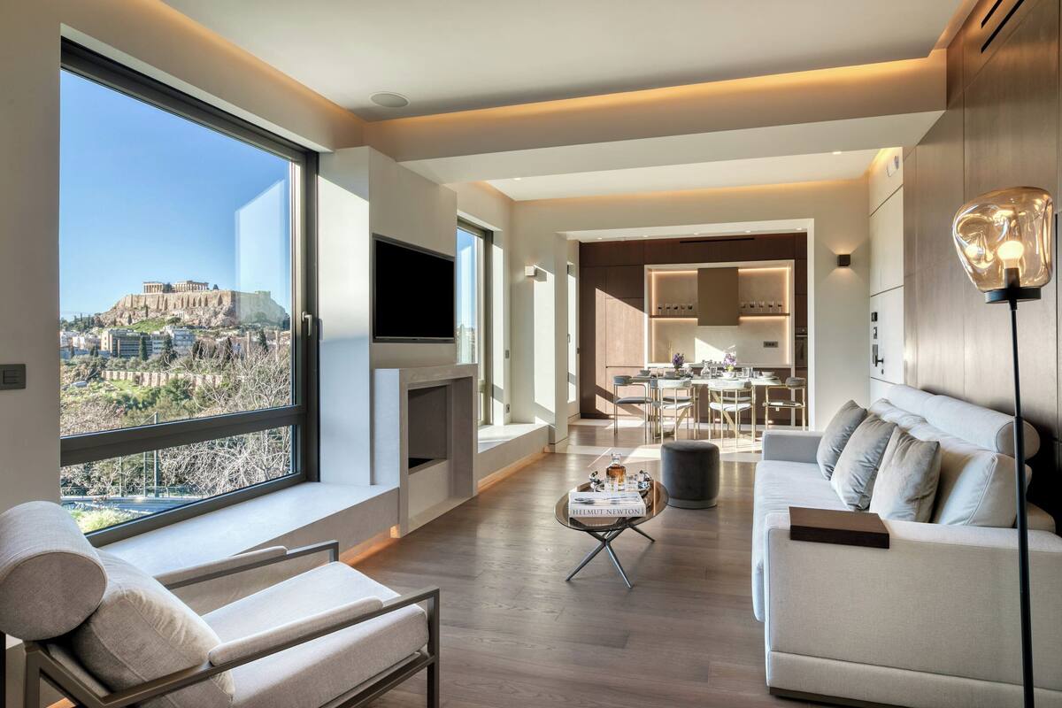 A modern living room with a large window overlooking a cityscape, a sofa, a coffee table, and a dining area in the background.